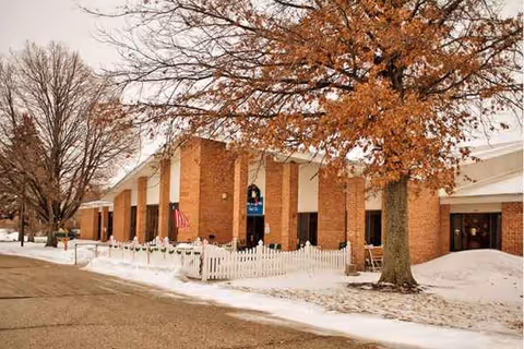 Snow blankets the sidewalk as a brick senior living facility entrance sits behind a white fence.