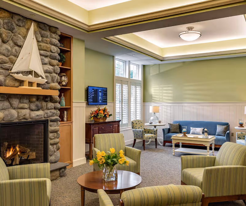 Bright, cozy lounge area with a stone fireplace, upholstered chairs and sofa arranged around coffee tables and floral arrangements.