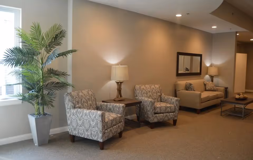 A cozy living room area with two patterned armchairs, a beige sofa with cushions, two table lamps on side tables, a rectangular mirror on the wall, a coffee table with decorative items, and a tall potted plant near a window.