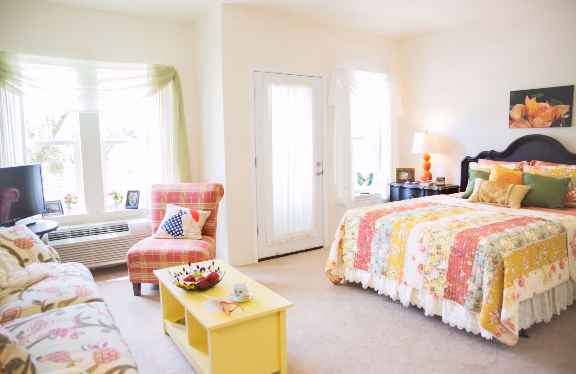 A bright and cozy bedroom with a large bed covered in a colorful patchwork quilt and multiple pillows. To the left, there is a plaid armchair with a decorative pillow, a floral-patterned sofa, and a yellow coffee table with a bowl of fruit and a teacup. The room has large windows with sheer curtains allowing natural light to fill the space, and a door with a sheer curtain. A bedside table with an orange lamp and framed pictures is next to the bed, and a floral painting hangs on the wall above the headboard.