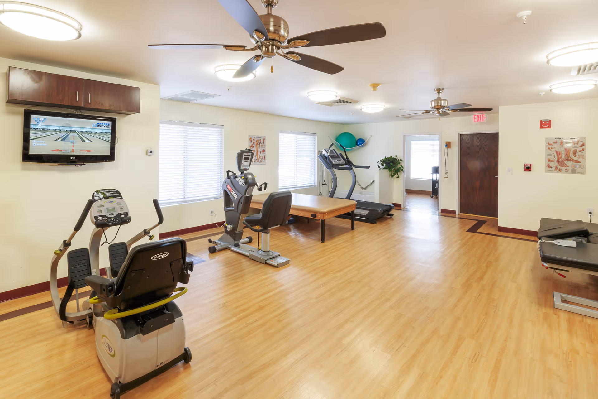 A rehabilitation center exercise room with wooden flooring, featuring exercise equipment including a recumbent bike, an upright bike, a treadmill, and a therapy table. The room has ceiling fans, bright overhead lights, windows with blinds, and a TV mounted on the wall displaying a bowling game. There are anatomical charts on the walls and some exercise balls on a shelf in the corner.