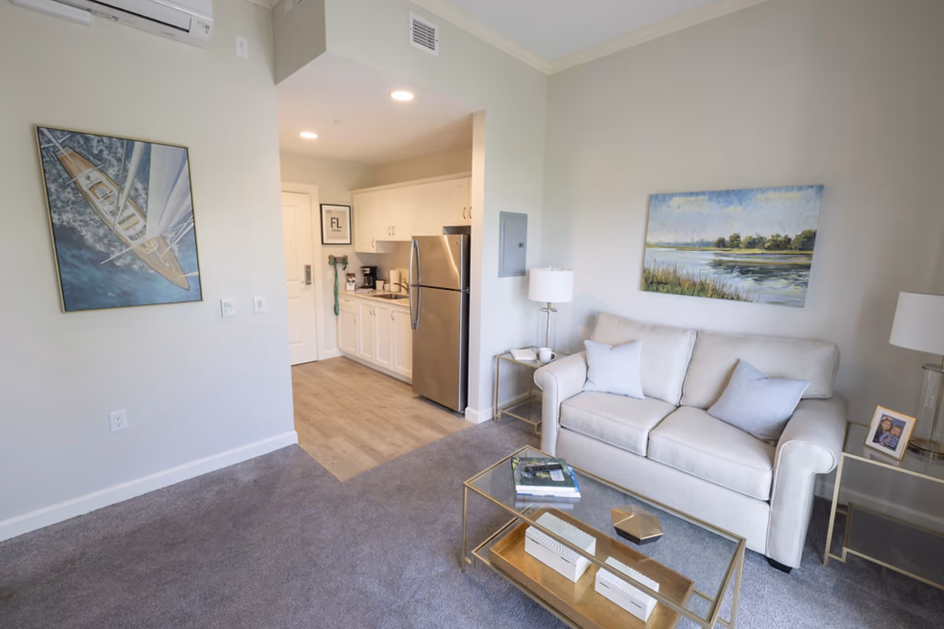 A cozy living room area with a beige sofa adorned with two light-colored pillows. A glass coffee table with gold trim holds books and decorative items. To the right of the sofa is a side table with a lamp and a framed photo. The room has light-colored walls with two paintings, one of a sailboat and another of a serene lakeside scene. In the background, a small kitchen area with white cabinets and a stainless steel refrigerator is visible.