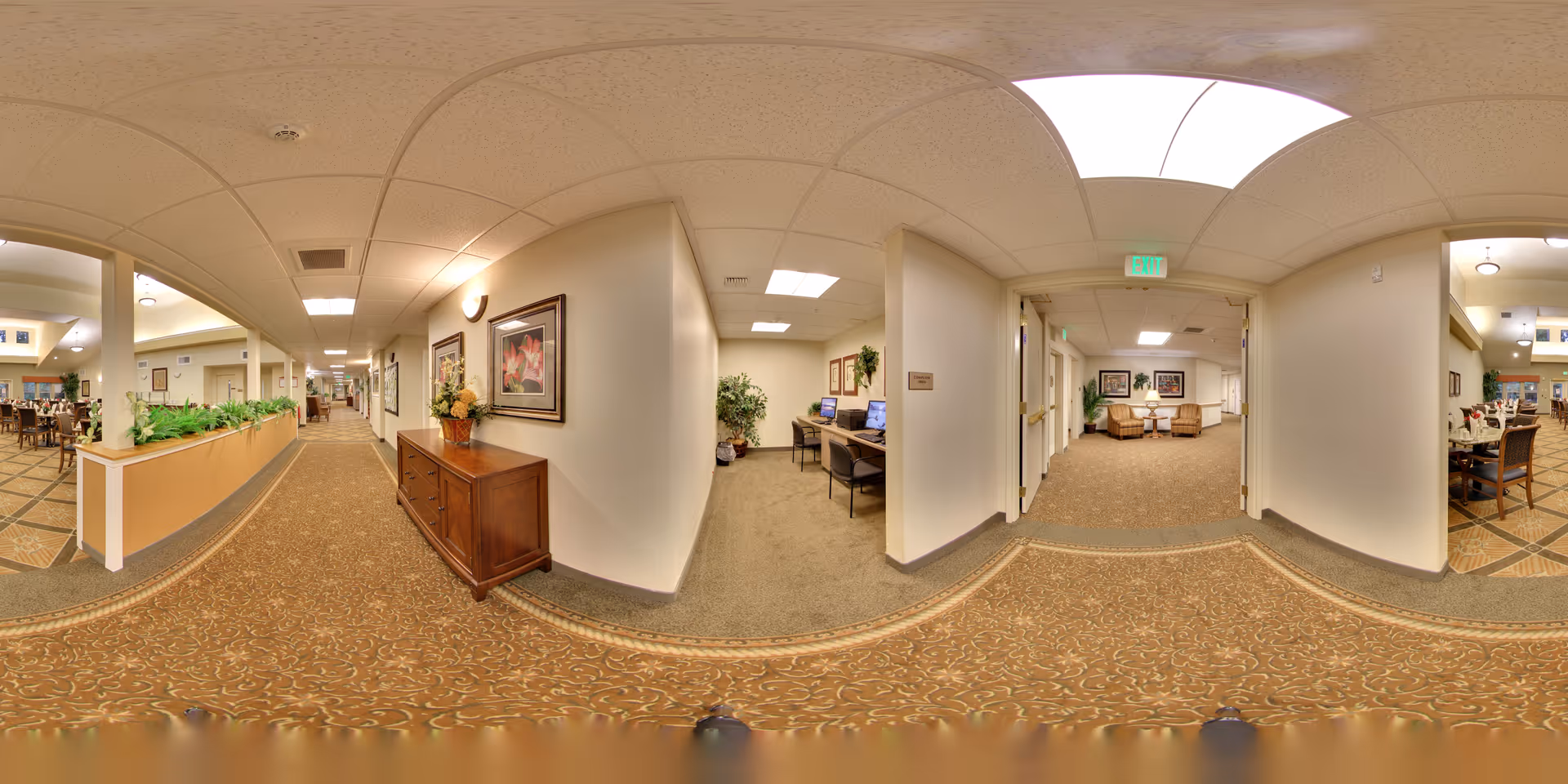 Carpeted interior hallway in a senior living facility with seating areas, framed artwork, a computer station, and adjacent dining spaces.