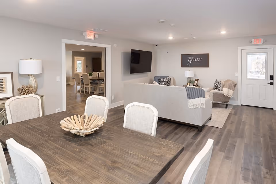 A modern senior living common area featuring a wooden dining table with four upholstered chairs in the foreground. Beyond the dining area is a living room with a gray sofa, armchair, a wall-mounted flat-screen TV, and a decorative sign on the wall. The room has light gray walls, wood flooring, and two exit doors with exit signs above them. A secondary dining area is visible through an open doorway.