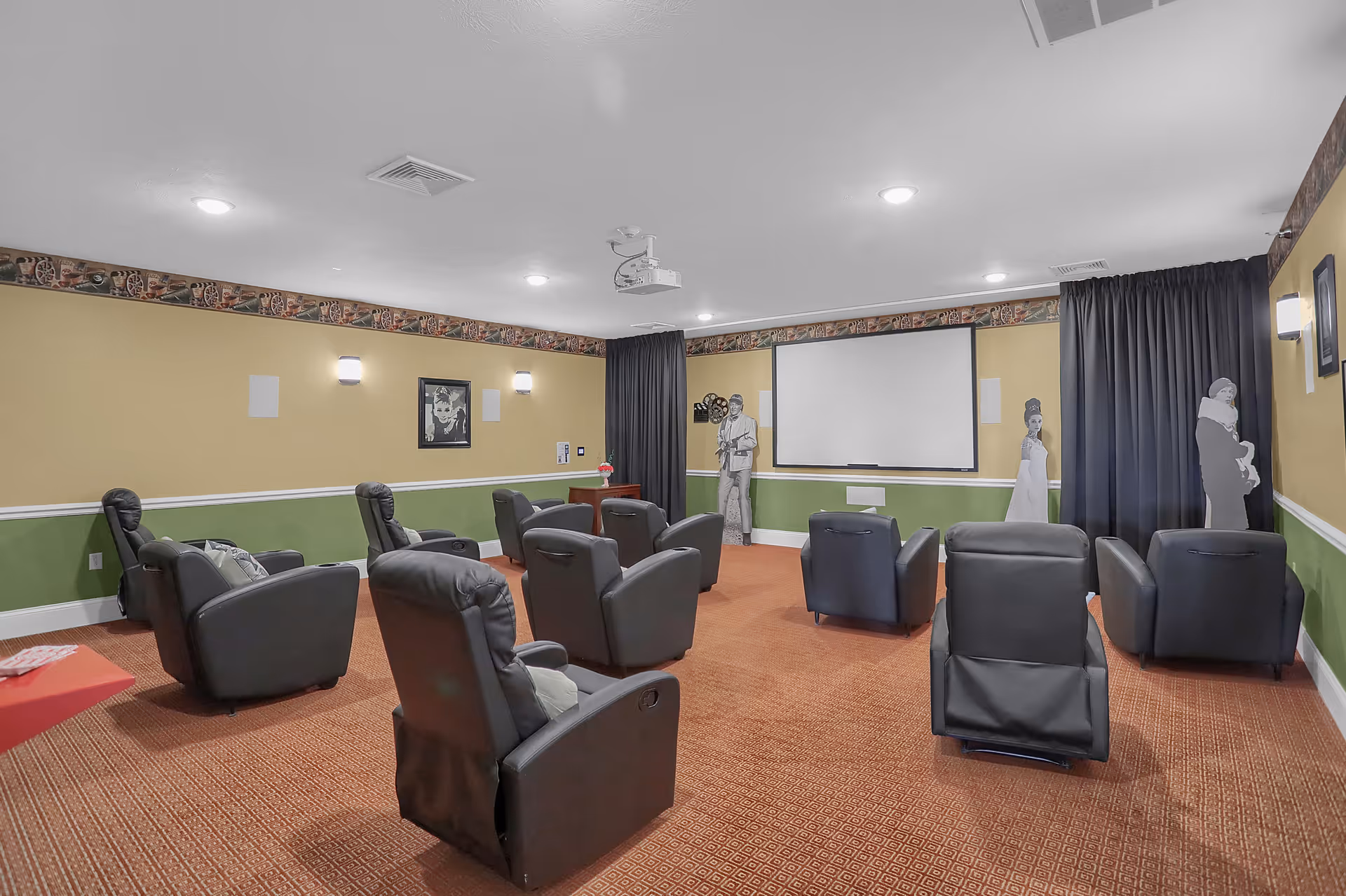 A small movie theater room with several black recliner chairs arranged in rows facing a large white projection screen. The walls are painted yellow and green with a decorative border near the ceiling. There are wall-mounted lights, framed artwork, and life-sized cutouts of classic movie characters near the screen. The floor is carpeted in a reddish pattern.