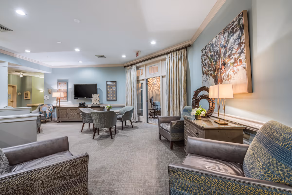 Comfortable senior living lounge with arranged armchairs, small tables, a wall-mounted TV, lamps, and large artwork.