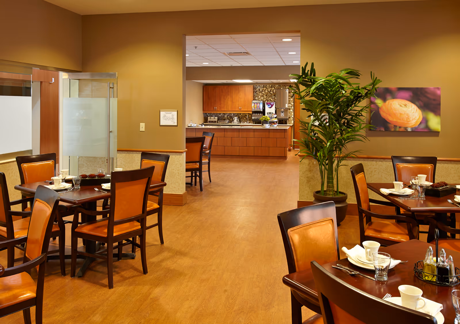 A warm and inviting dining room with wooden tables and chairs featuring orange cushions. The tables are set with white cups, glasses, and napkins. In the background, there is a kitchen area with wooden cabinets and a coffee machine. A large green potted plant and a colorful flower painting adorn the walls.