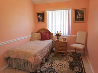 A small bedroom with peach-colored walls, a single bed with floral bedding and pillows, a wooden nightstand with a vase of flowers and a small decorative item, a white chair, two framed floral paintings on the walls, and a window with vertical blinds.