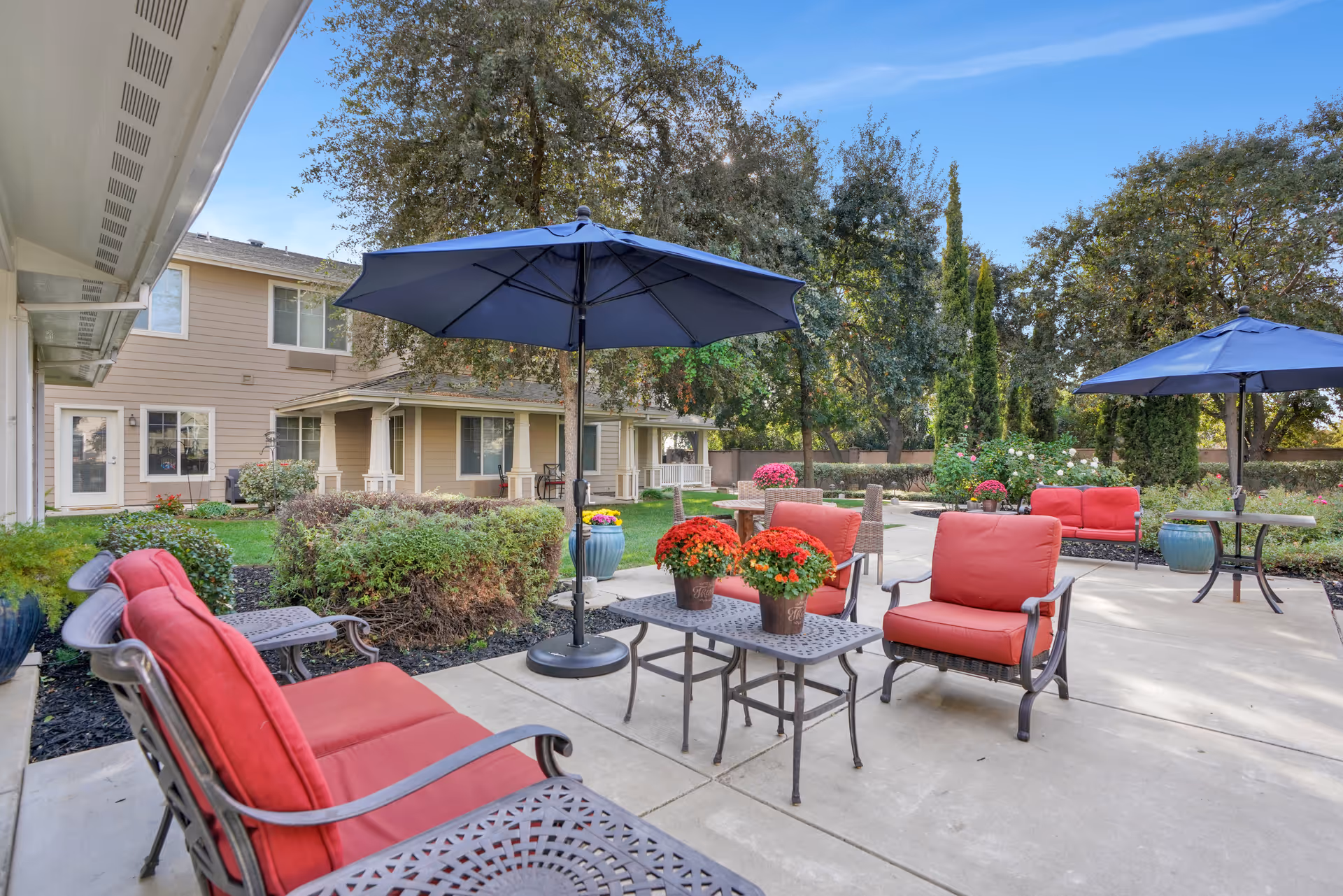 Outdoor patio area at The Commons At Elk Grove with red cushioned chairs and sofas, black metal tables, blue umbrellas, potted flowers, and surrounding greenery including trees and bushes under a clear blue sky.
