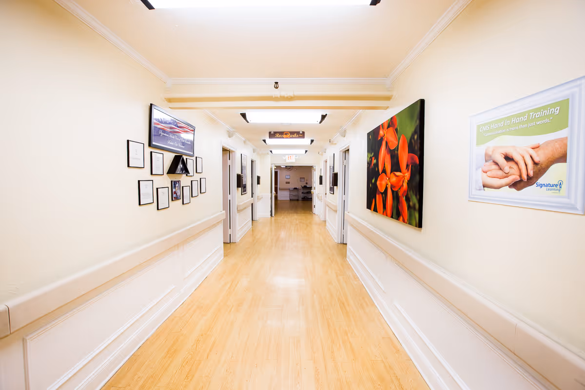 A well-lit hallway in a senior living facility with light-colored walls and wooden flooring. The walls are decorated with framed pictures and certificates, including a photo of red flowers and a poster about CMS Hand in Hand Training. Several doorways line the hallway, and there is a sign hanging from the ceiling further down the corridor.