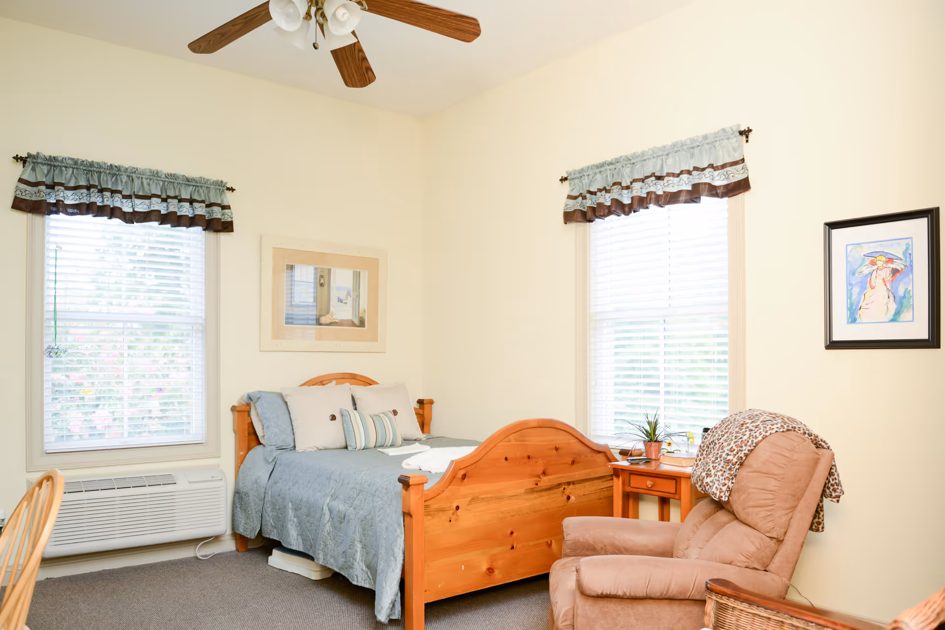 A cozy bedroom with a wooden bed frame made of light-colored wood, dressed in blue bedding and several pillows. There are two windows with white blinds and blue valances with brown trim. A ceiling fan with wooden blades and light fixtures is mounted on the ceiling. Next to the bed is a wooden nightstand with a small plant and a lamp. A comfortable beige recliner chair with a leopard print throw is positioned near the nightstand. The walls are painted a soft cream color and decorated with two framed pictures.