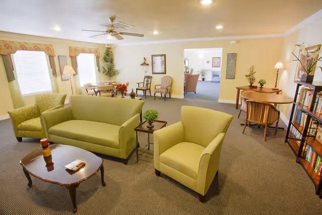 Well-lit communal lounge with green sofas and armchairs, coffee tables, bookshelves, and small dining tables.