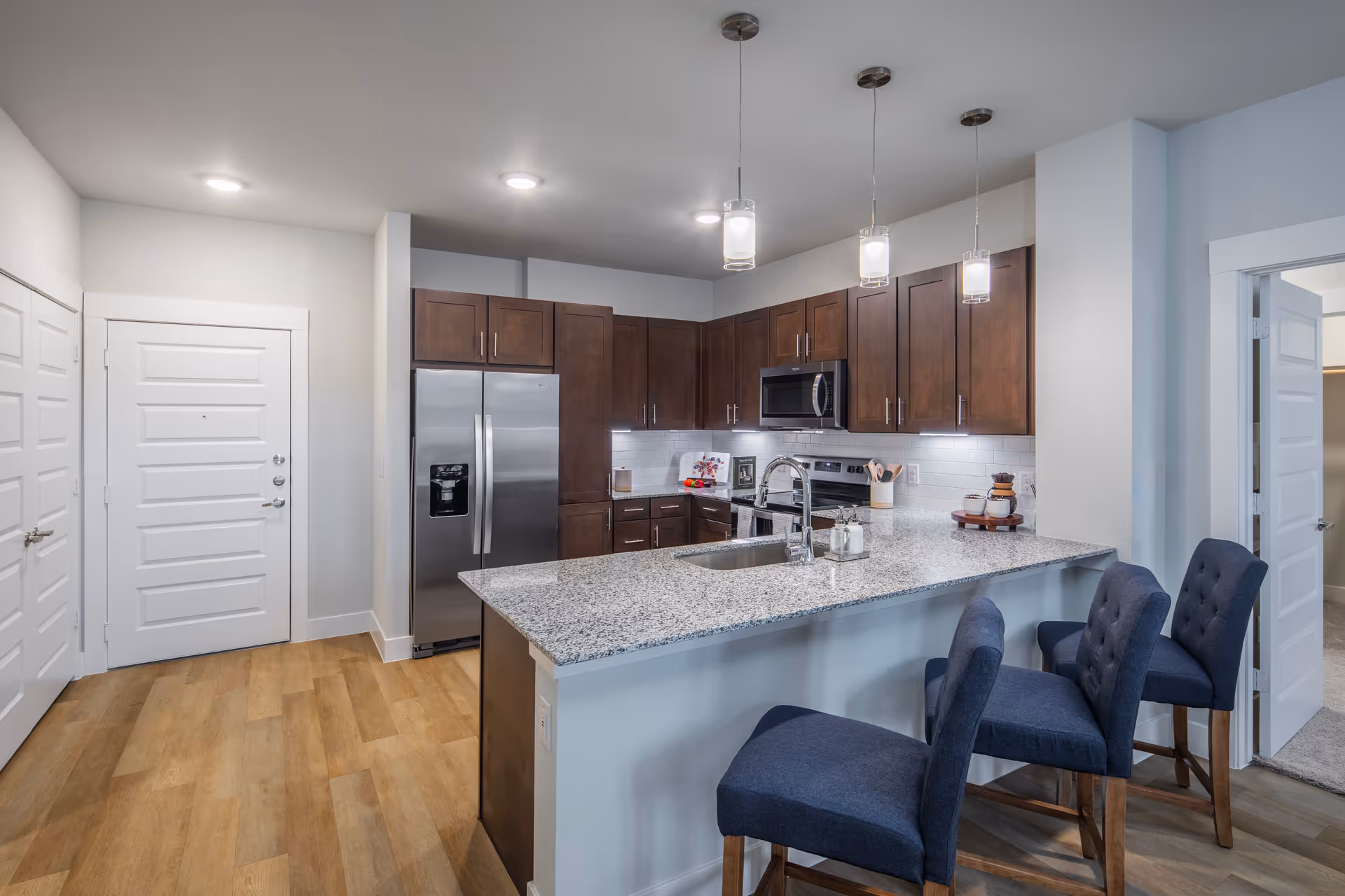 Modern kitchen with granite countertop island featuring a sink and three blue upholstered bar stools. The kitchen has dark wooden cabinets, stainless steel refrigerator, microwave, and stove. Pendant lights hang above the island, and there is wood flooring throughout.
