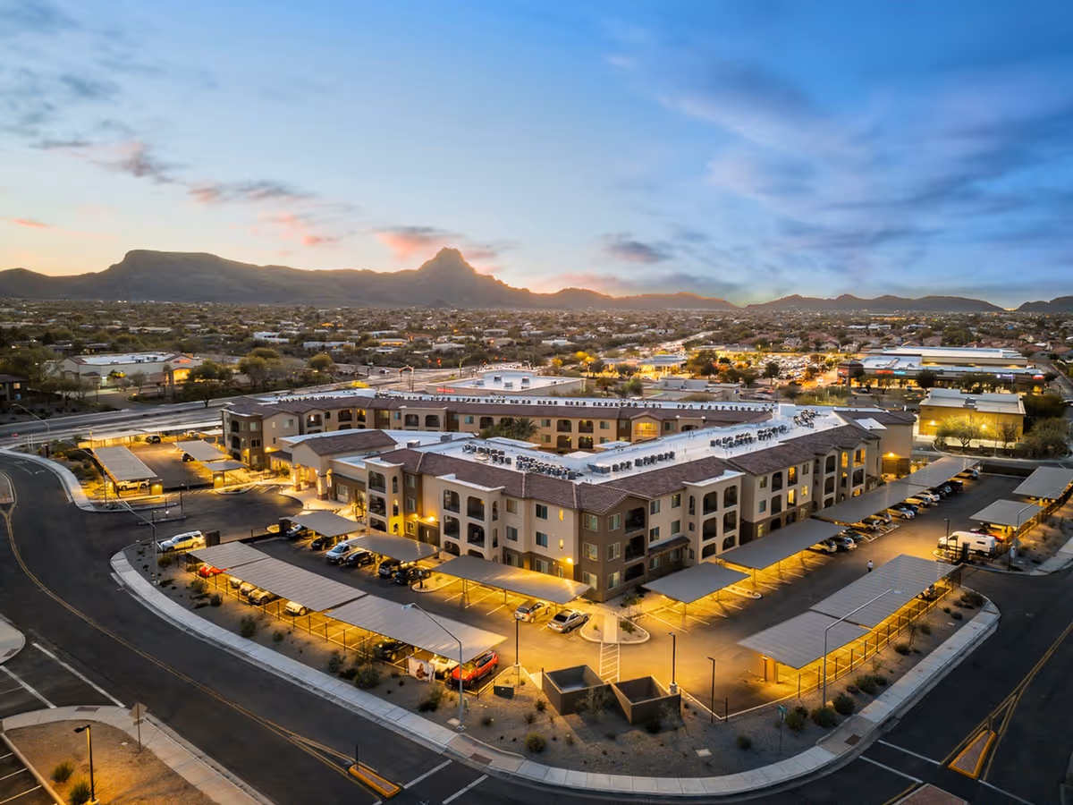 Aerial view of Album Marana facility at dusk, showing a large, multi-story building with covered parking areas surrounding it. The building is illuminated with warm lights and is set against a backdrop of mountains and a partly cloudy sky.