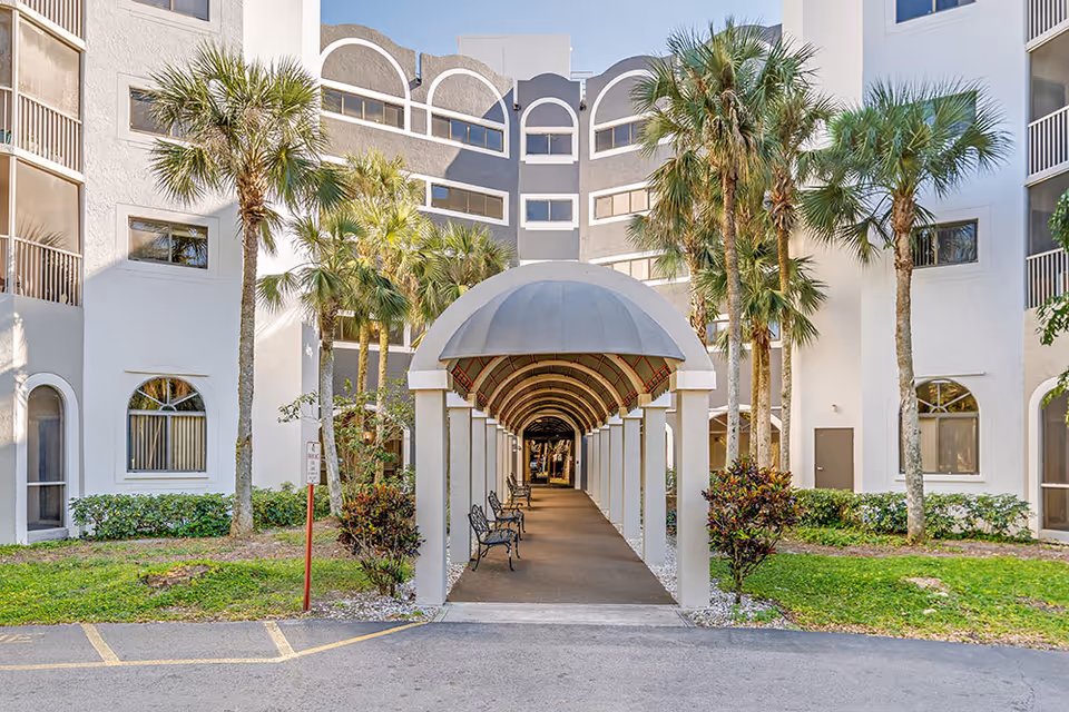 Exterior view of Forest Trace Senior Living showing a covered walkway with benches leading into the building, surrounded by palm trees and greenery.