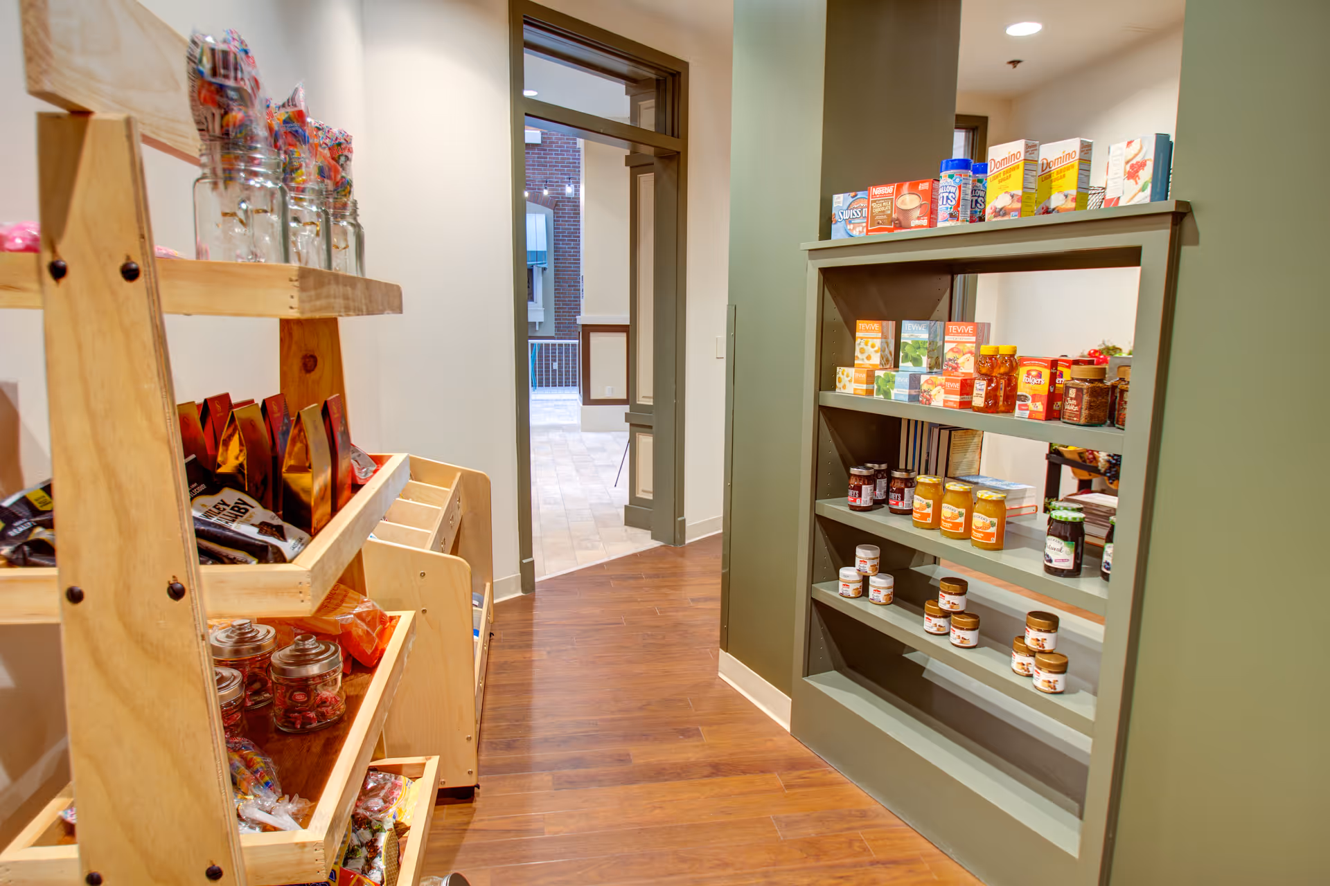 A small pantry area with wooden shelves stocked with various packaged food items, jars, and candy. The floor is wooden, and there is an open doorway leading to a tiled area with a window showing an outdoor view.
