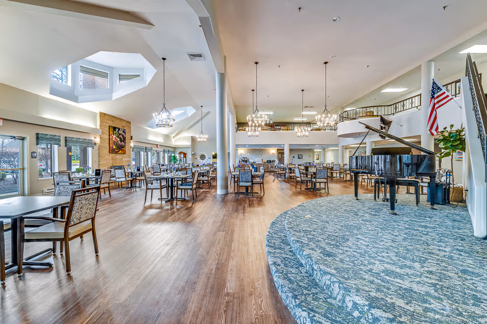 Spacious dining/activity room with multiple tables and chairs, a raised stage with a grand piano, chandeliers, and large skylights.