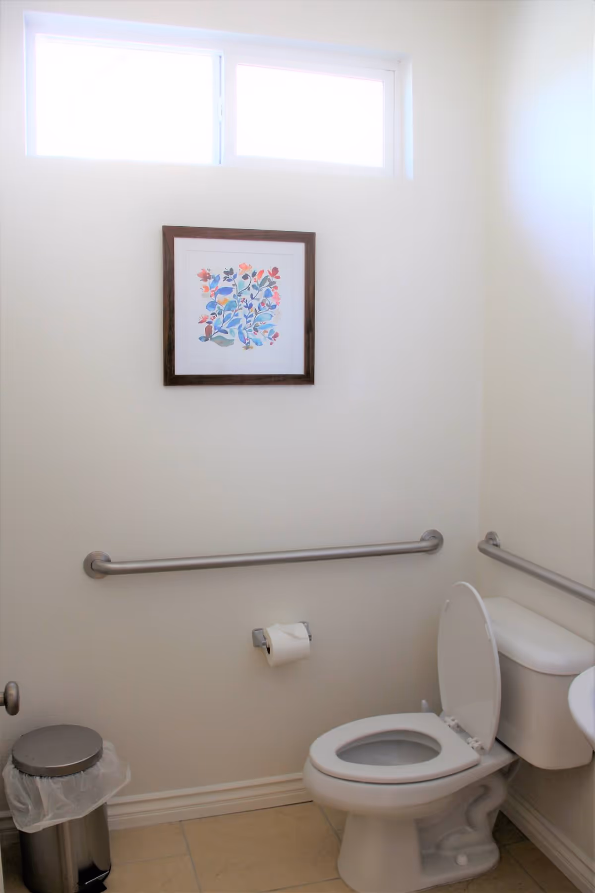 A clean bathroom with a white toilet with the lid open, a metal trash can with a plastic liner, beige tiled floor, white walls, a horizontal grab bar on the wall behind the toilet, and a framed colorful floral artwork hanging above the grab bar. There is a narrow horizontal window near the ceiling letting in natural light.