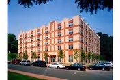 A multi-story brick and beige building with numerous windows, surrounded by parked cars and greenery under a clear blue sky.
