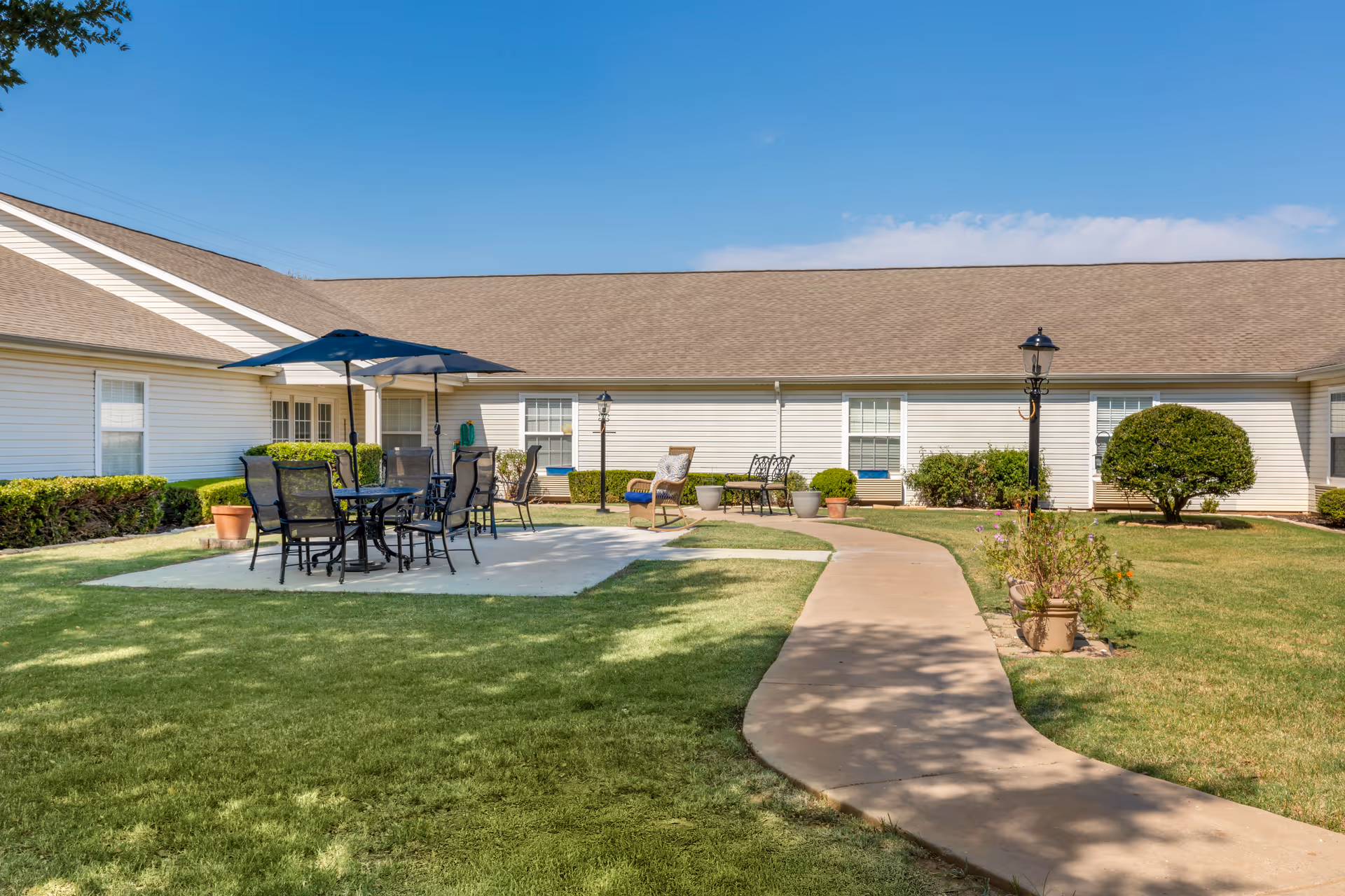 Outdoor patio area at Brookdale Midwestern featuring a concrete pathway leading to a seating area with a table, chairs, and umbrellas. The area is surrounded by green grass, potted plants, bushes, and a building with white siding and multiple windows under a clear blue sky.