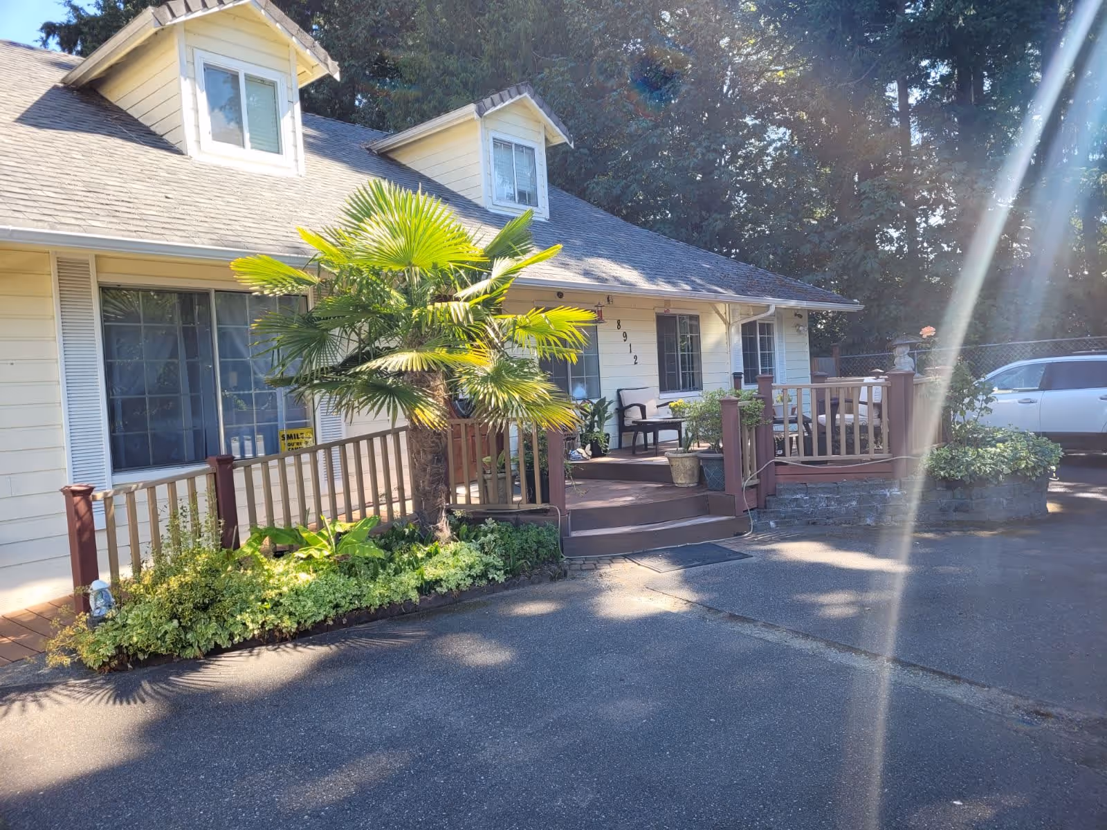 Exterior view of a single-story residential building with light yellow siding and a gray shingled roof. The entrance has a small wooden porch with steps, potted plants, and outdoor seating. There is a palm tree and other greenery in front of the porch. A white vehicle is parked to the right side of the building. Sunlight creates a lens flare effect in the image.