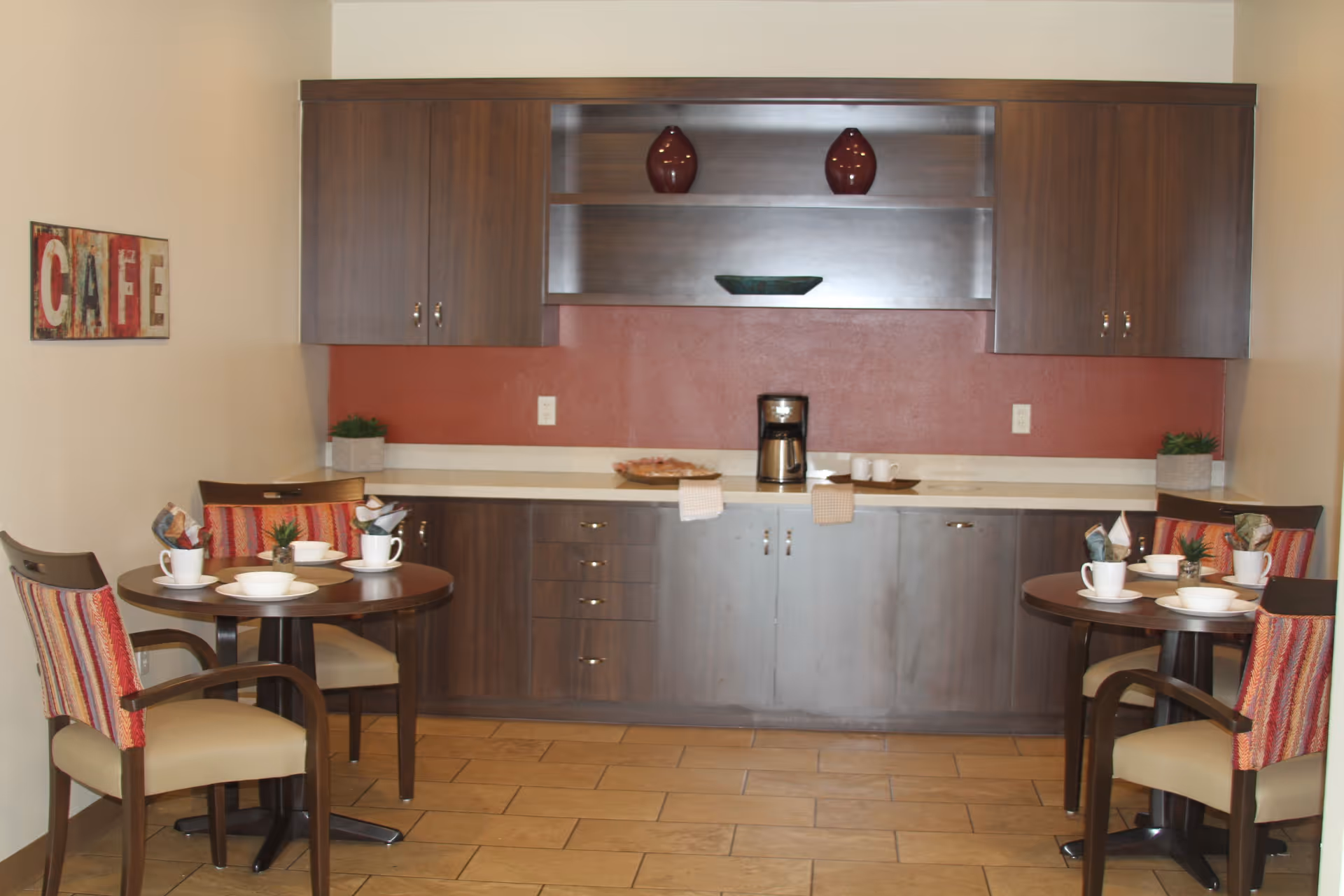 Small dining area with two round tables set for meals in front of a kitchenette with wood cabinets and a coffee maker.