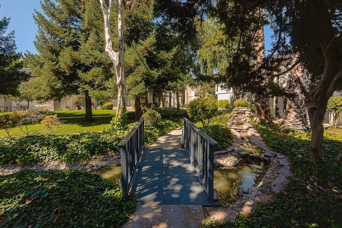 A small blue wooden bridge over a narrow stream with rocks and greenery on both sides, surrounded by lush trees and bushes in a garden area of a retirement community. Buildings are visible in the background under a clear blue sky.