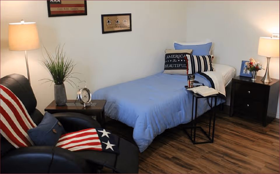 A cozy bedroom with a single bed dressed in blue bedding and patriotic-themed pillows. Next to the bed is a small black nightstand with a lamp, a framed photo, and a small flower arrangement. A black recliner chair with an American flag-themed blanket and pillow is positioned near a wooden side table holding a lamp, a plant, and a clock. The room has wooden flooring and white walls with framed patriotic artwork.