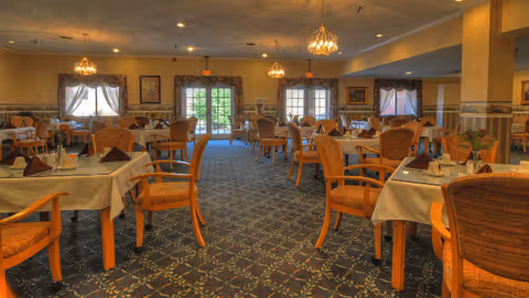 Large dining room with neatly set tables and wicker-backed chairs, chandeliers, patterned carpet, and double doors at the far end.