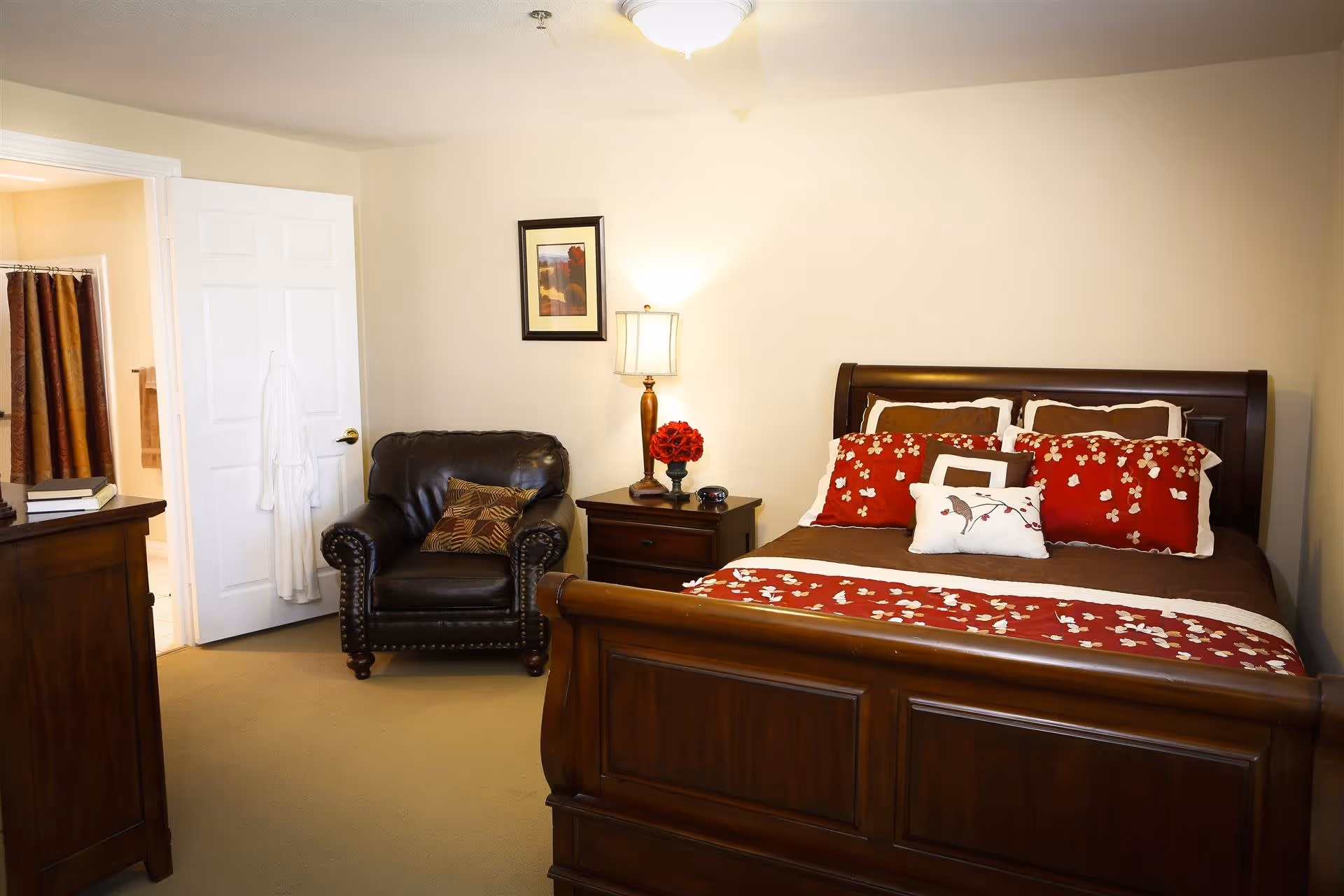 A cozy bedroom with a wooden bed featuring red and brown bedding with floral patterns. Next to the bed is a wooden nightstand with a lamp and a vase of red flowers. There is a dark leather armchair with a patterned pillow, a framed picture on the wall, and an open door leading to a bathroom with a shower curtain and a white robe hanging on the door.