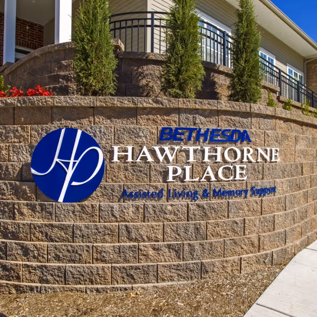 Stone wall with the Bethesda Hawthorne Place logo and text reading 'Assisted Living & Memory Support' in front of a building with small trees planted on top of the wall.