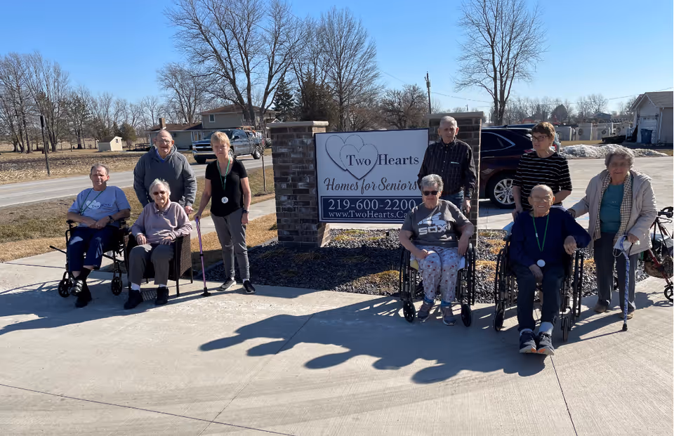 A group of senior residents and staff posing outside next to a brick sign that reads "Two Hearts Homes for Seniors."