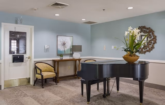 Interior room with a black grand piano in the foreground, a decorative round mirror on the wall, a table with a lamp and a small plant, and a yellow upholstered chair near a white door with a window.