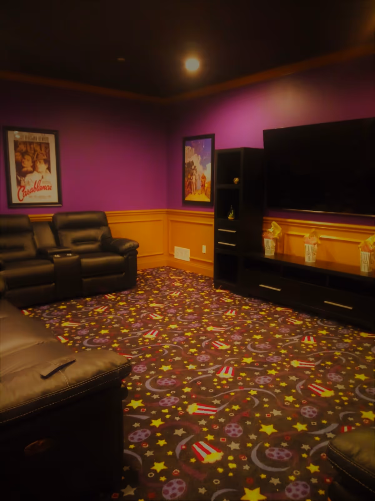 A cozy entertainment room with purple walls and wooden paneling. The room features black leather recliner chairs, a large flat-screen TV mounted on the wall, and a black entertainment unit with decorative popcorn containers. The carpet has a colorful pattern with stars, moons, and popcorn motifs. Two framed movie posters are hung on the walls.