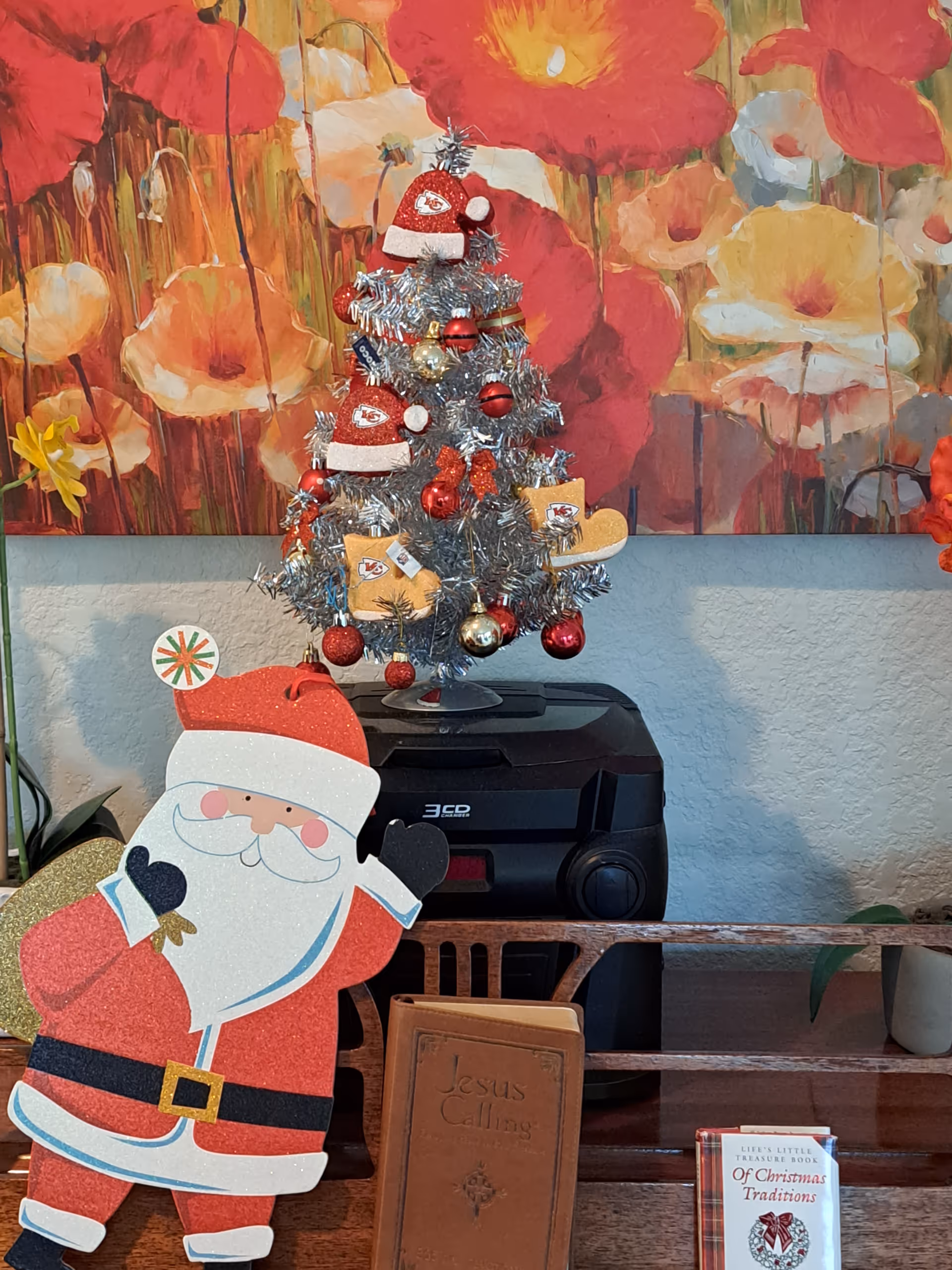 A festive holiday display featuring a small silver Christmas tree decorated with red and gold ornaments and Kansas City Chiefs-themed decorations, including mini hats and mittens. In front of the tree is a cheerful Santa Claus cutout. Below the display are two books, one titled 'Jesus Calling' and the other 'Luci's Little Treasure Book Of Christmas Traditions.' The background includes a colorful floral painting with large red, orange, and yellow flowers.