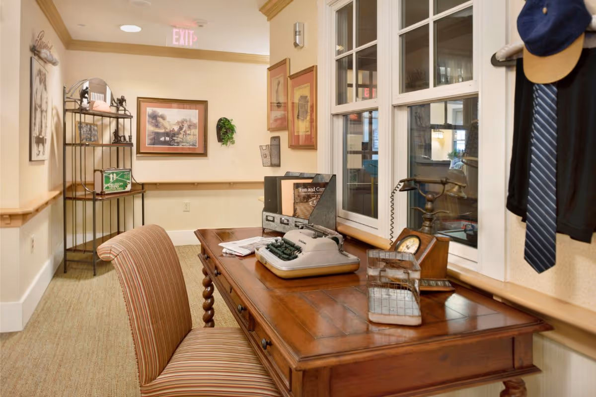 A cozy interior space featuring a wooden desk with a vintage typewriter, an old-fashioned telephone, and a wire letter holder. A striped upholstered chair is positioned in front of the desk. The wall behind the desk has windows with white frames, and there are framed pictures and a small plant on the adjacent wall. A metal shelving unit with decorative items is visible in the background, along with an exit sign on the ceiling.