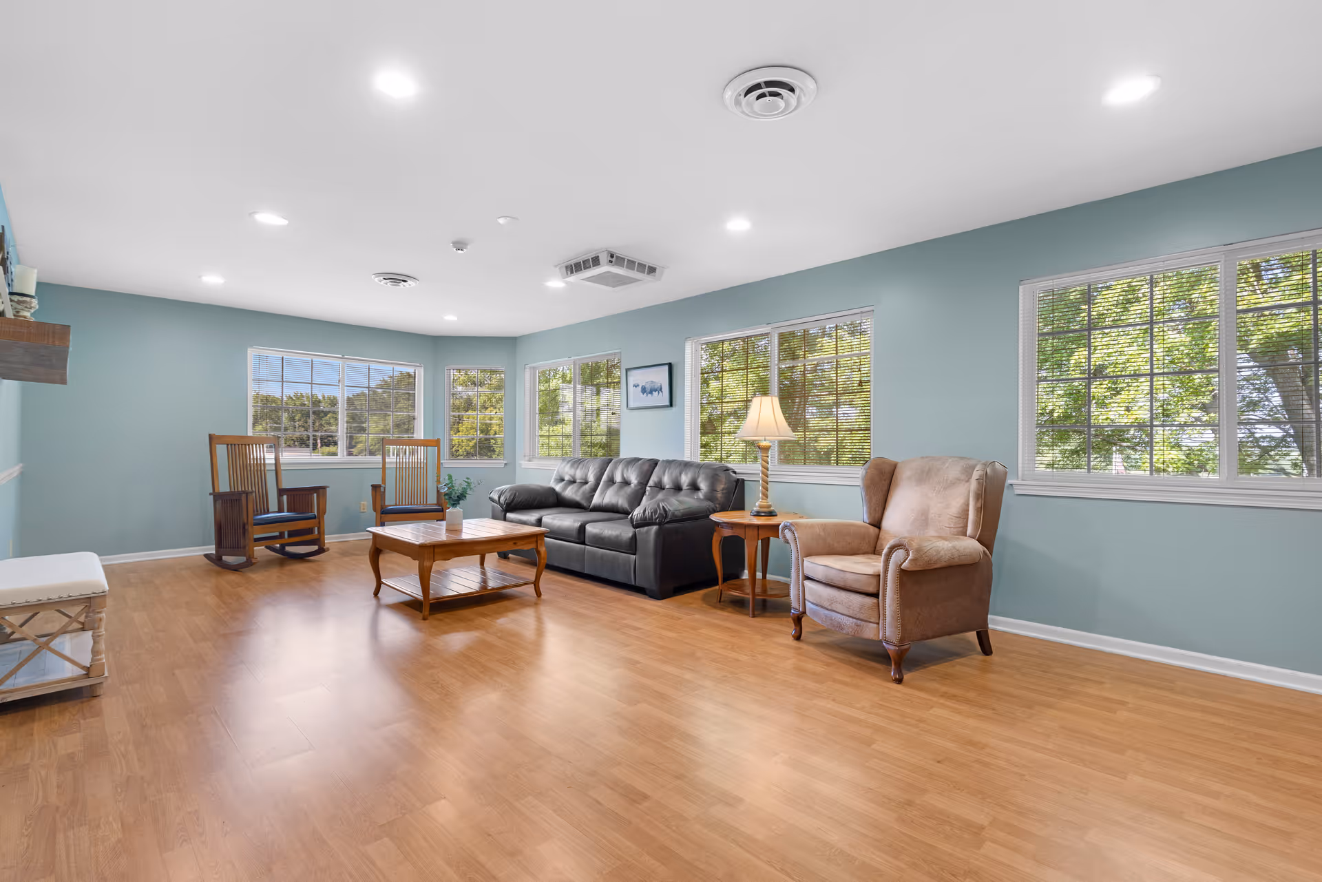 A bright and spacious living room with light blue walls and large windows letting in natural light. The room features a black leather sofa, a beige armchair, a wooden coffee table, two wooden rocking chairs, a side table with a lamp, and hardwood flooring.
