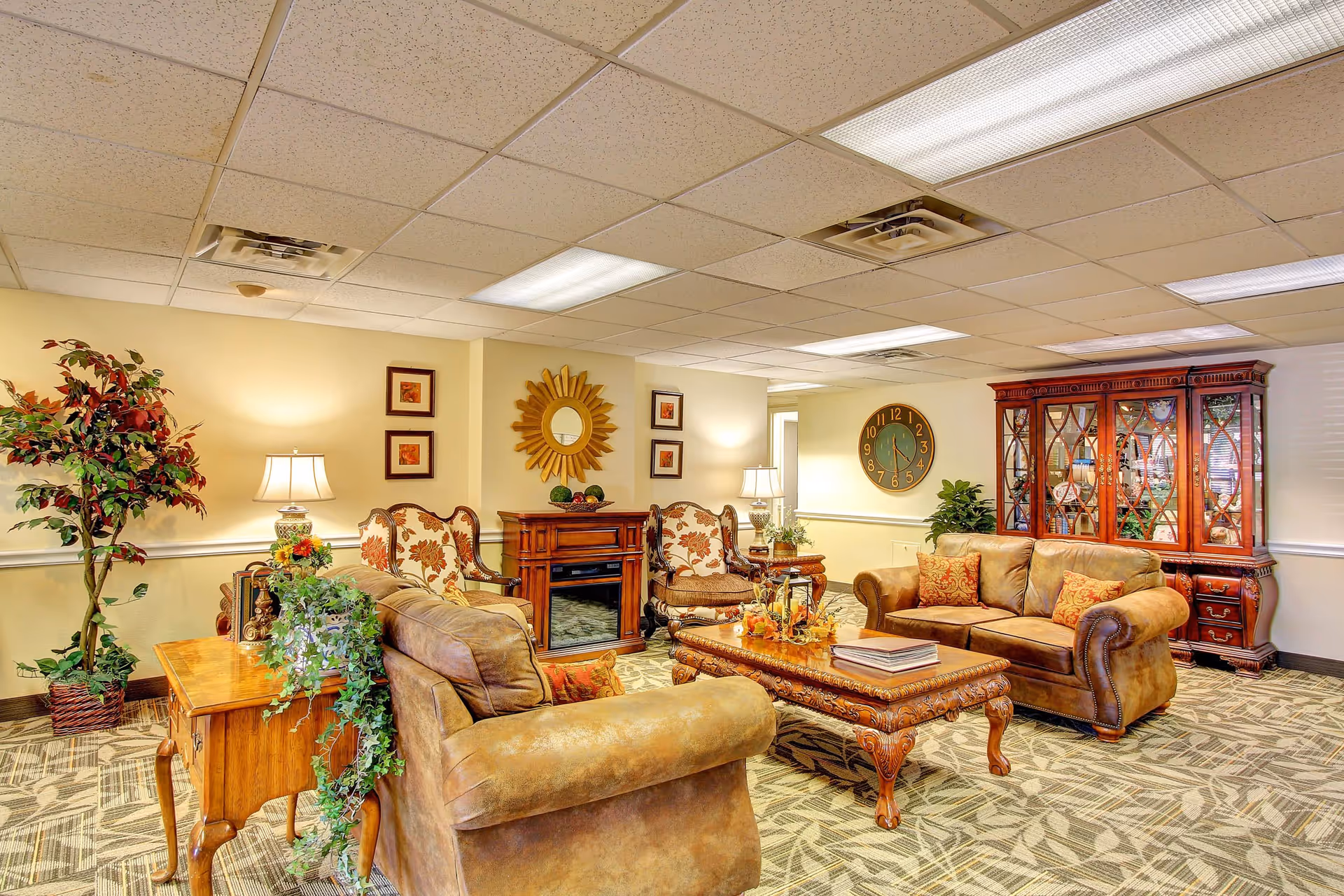 A cozy living room area in a senior living facility featuring two brown leather sofas, two floral upholstered armchairs, a wooden coffee table with decorative items, a wooden side table with a lamp and plants, a large wooden cabinet with glass doors, a wall clock, and a decorative mirror on the wall. The room has patterned carpet flooring and a drop ceiling with fluorescent lighting.