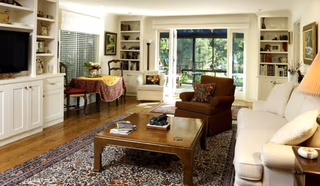 Bright living room with a sofa and armchair, wooden coffee table on a patterned rug, built-in white shelving, and sliding glass doors looking out to greenery.