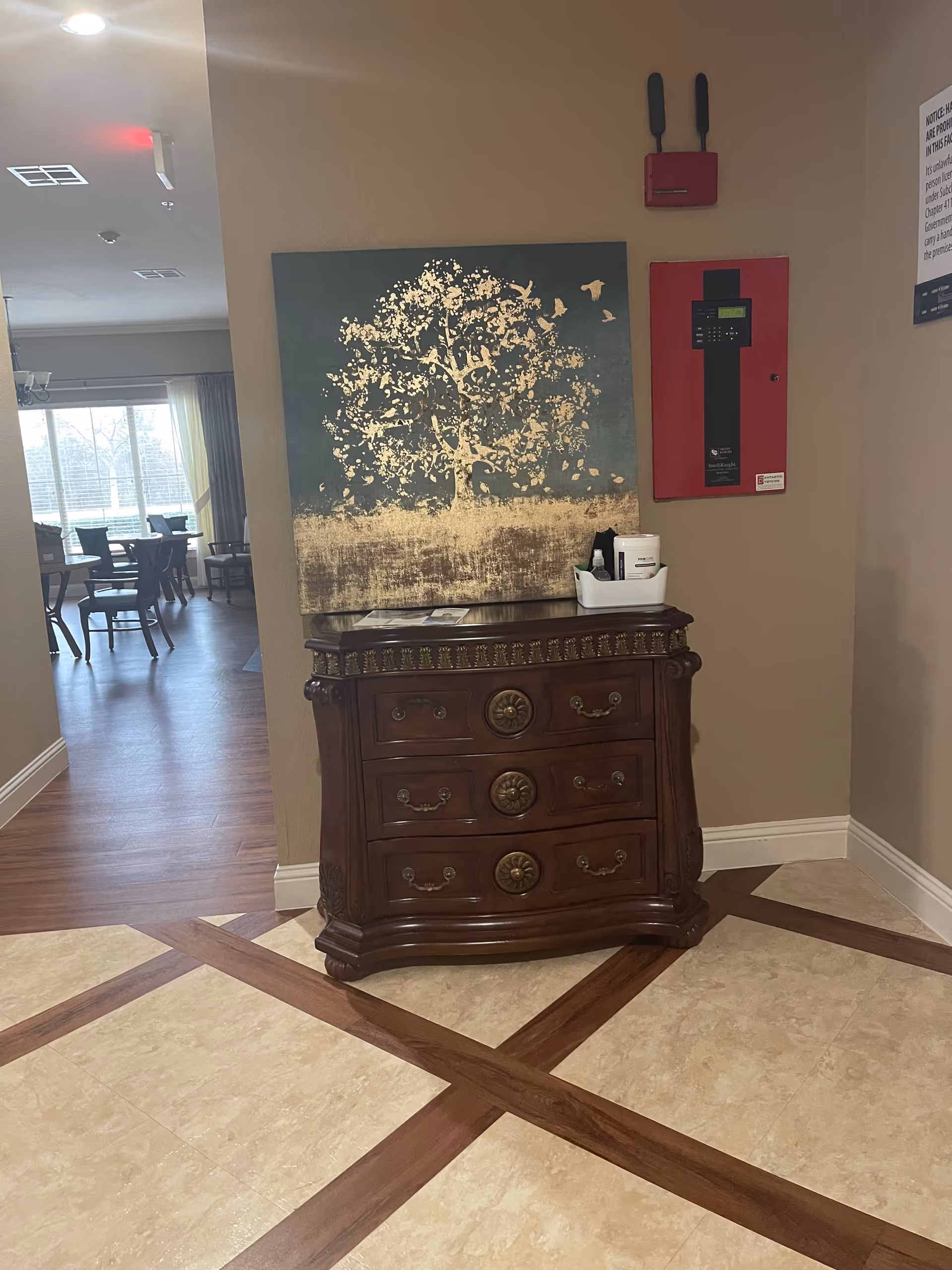 A corner of an interior space featuring a wooden chest of drawers with ornate handles and decorative carvings. Above the chest is a large painting of a tree with gold leaves and birds on a dark background. To the right of the painting is a red fire alarm control panel mounted on the wall. The floor has a tile and wood pattern, and in the background, there is a dining area with tables and chairs near large windows with curtains.