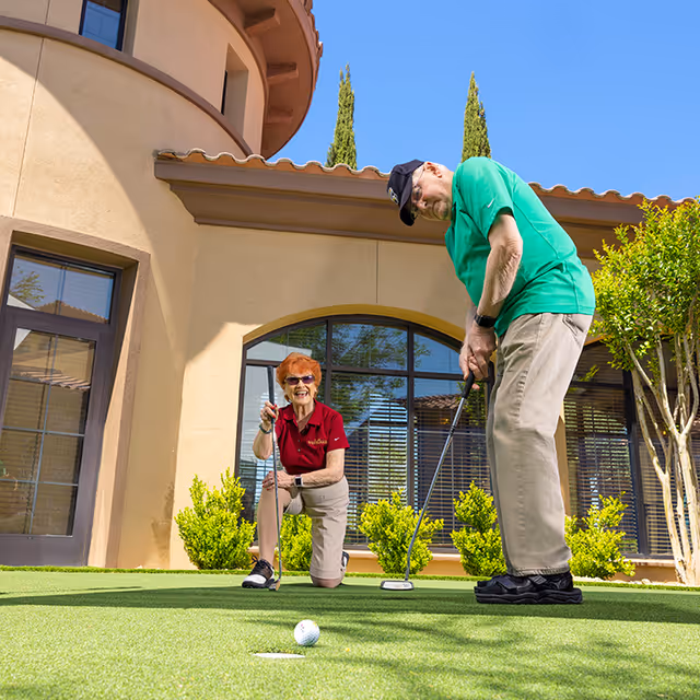 An elderly man and woman playing mini golf on a putting green outside a building with large windows and beige stucco walls. The man is standing and preparing to putt, while the woman is kneeling and smiling, holding a golf club. The sky is clear and blue, and there are green bushes and trees around the area.