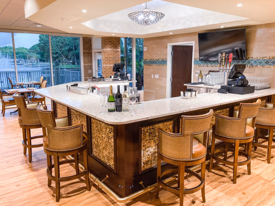 Interior view of a bar area with a large U-shaped counter surrounded by wooden bar stools with cushioned seats. Bottles of wine and glasses are placed on the counter. Behind the bar, there is a mounted flat-screen TV and beer taps. Large windows offer a view of a lake with trees and a fountain outside. The floor is wooden, and the walls have decorative tile accents.