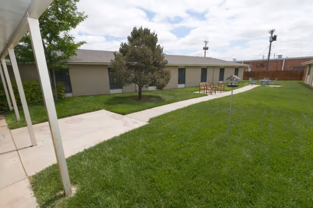 Grassy courtyard with a concrete path, outdoor chairs, a small tree, and a single-story building under a partly cloudy sky.