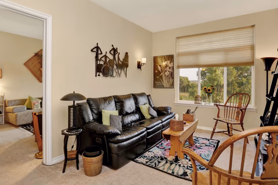A cozy living room with a black leather sofa adorned with green and checkered pillows. A wooden coffee table sits on a floral rug in front of the sofa. There are two wooden chairs, one near the window and another partially visible in the foreground. The window has a beige blind and offers a view of green trees outside. The walls are decorated with a metal wall art piece and a framed picture. A black side table with a lamp and a wicker wastebasket are next to the sofa. Another room with a gray armchair and a lamp is visible through an open doorway.