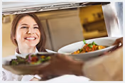 A smiling chef in a kitchen handing plates of food through a serving window to another person.