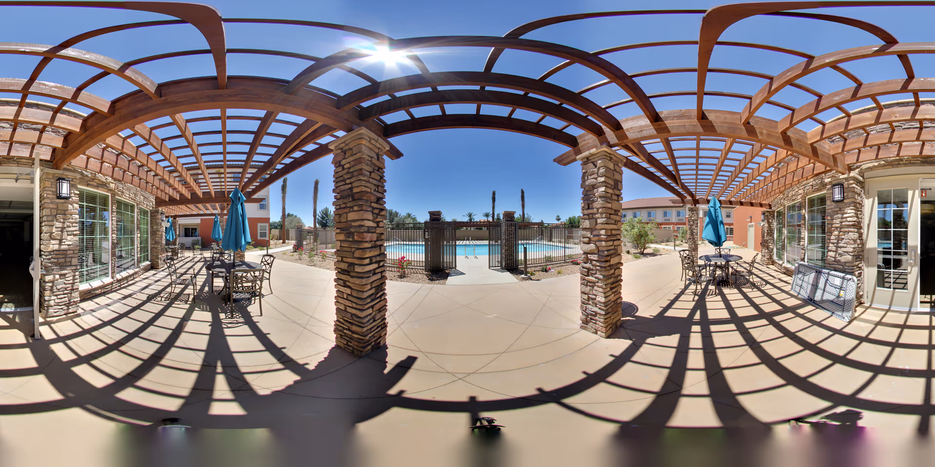 Shaded outdoor patio with a wooden pergola and stone columns overlooking a gated swimming pool with tables and umbrellas.
