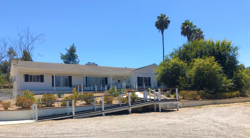 Single-story white building with a ramp leading to the entrance, surrounded by bushes and trees including two tall palm trees under a clear blue sky.