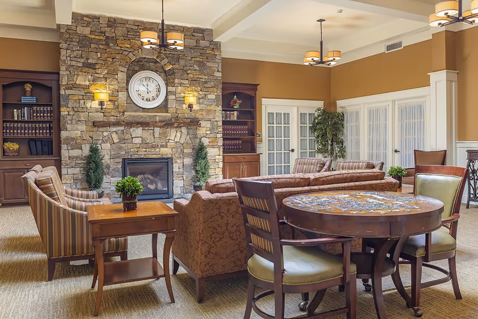 A cozy living room area with a stone fireplace and a clock above it. The room features comfortable upholstered chairs and sofas arranged around the fireplace, wooden bookshelves filled with books, a round wooden table with chairs, and warm lighting fixtures on the ceiling and walls. There are also some potted plants and French doors with glass panes in the background.