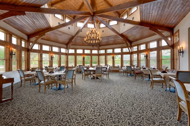 Spacious senior living dining room with multiple tables and chairs under a vaulted wooden ceiling and large windows.