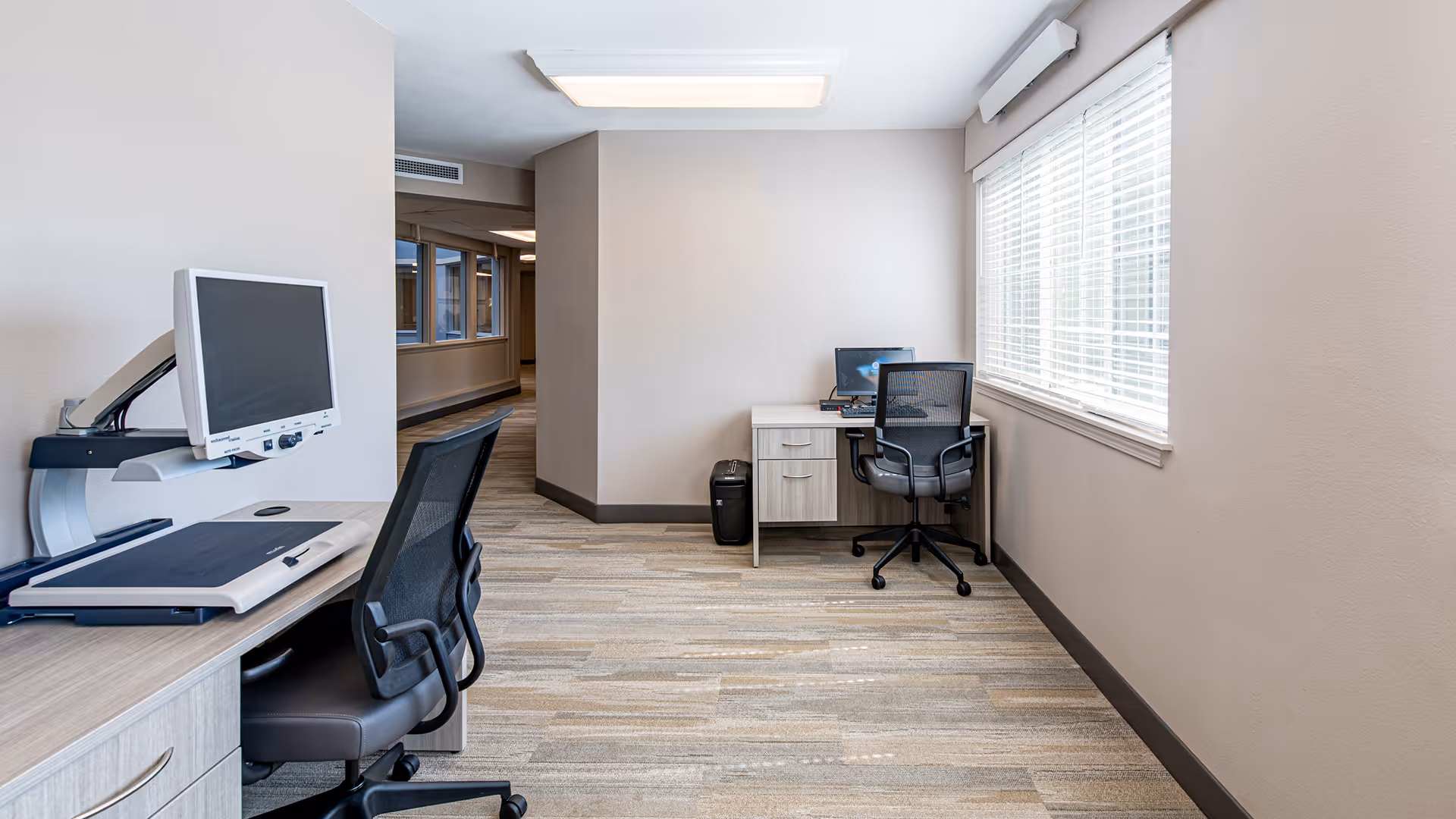 Small, bright office/computer room with two desks, office chairs, a computer monitor, and a large window with blinds.
